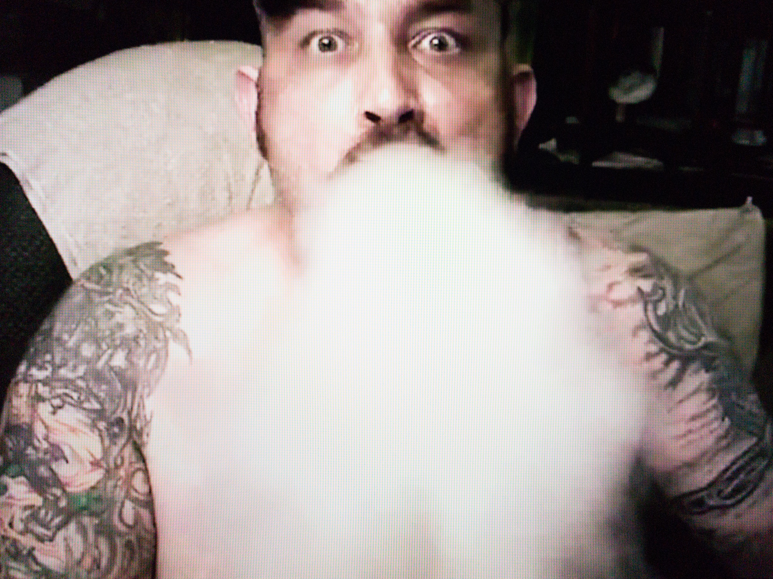 A bare-chested, bearded man with tattoos on arms and shoulders is looking straight at us with both eyes wide open, while exhaling a large volume of smoke that covers his entire chest.