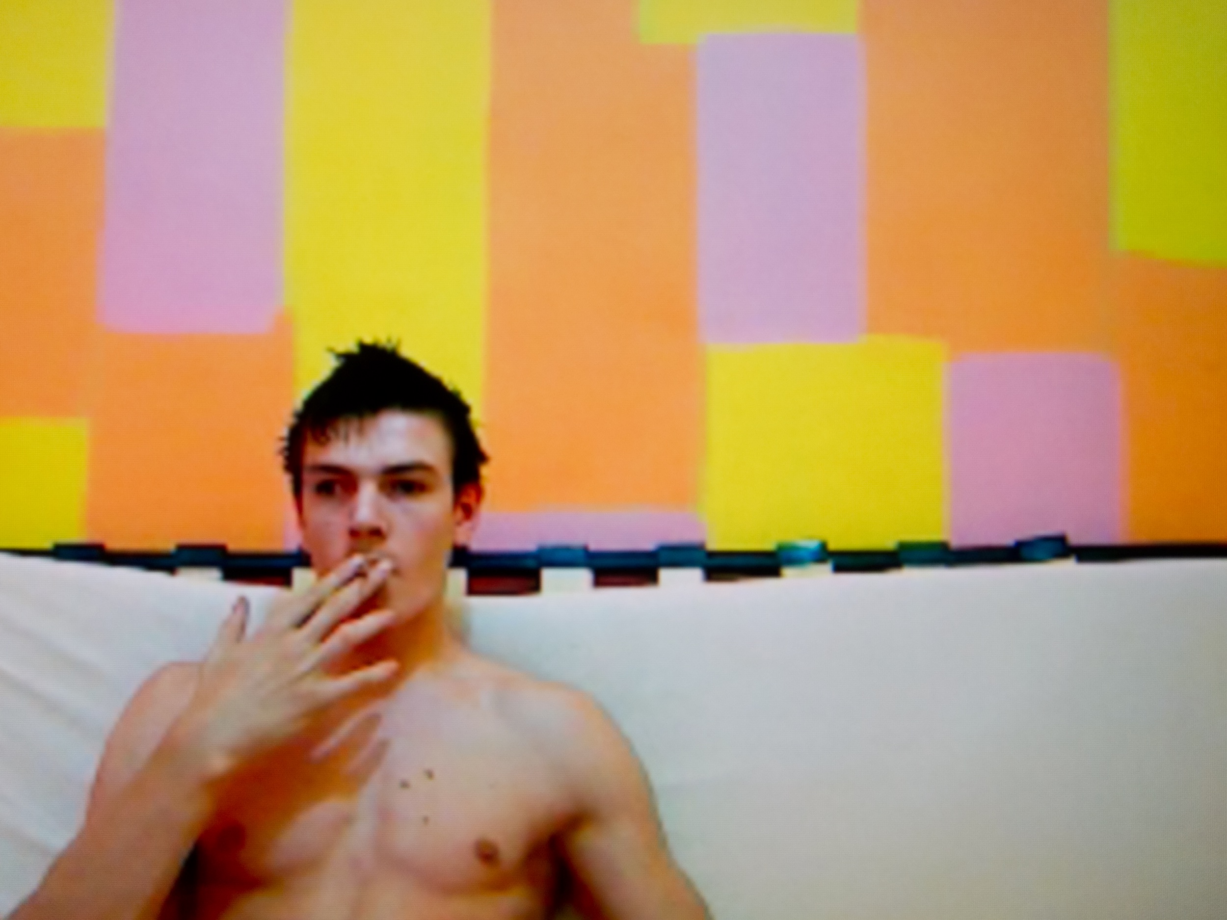 A bare-chested male stripper takes a drag on a cigarette while sitting on a white sofa in front of a wall painted with yellow, orange, and lavender rectangular shapes