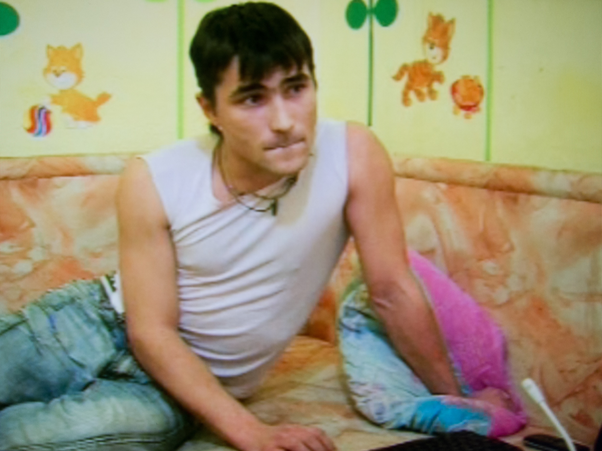 A male stripper in white tanktop sits on an orange marbled sofa in front of a green wall with cat cartoons