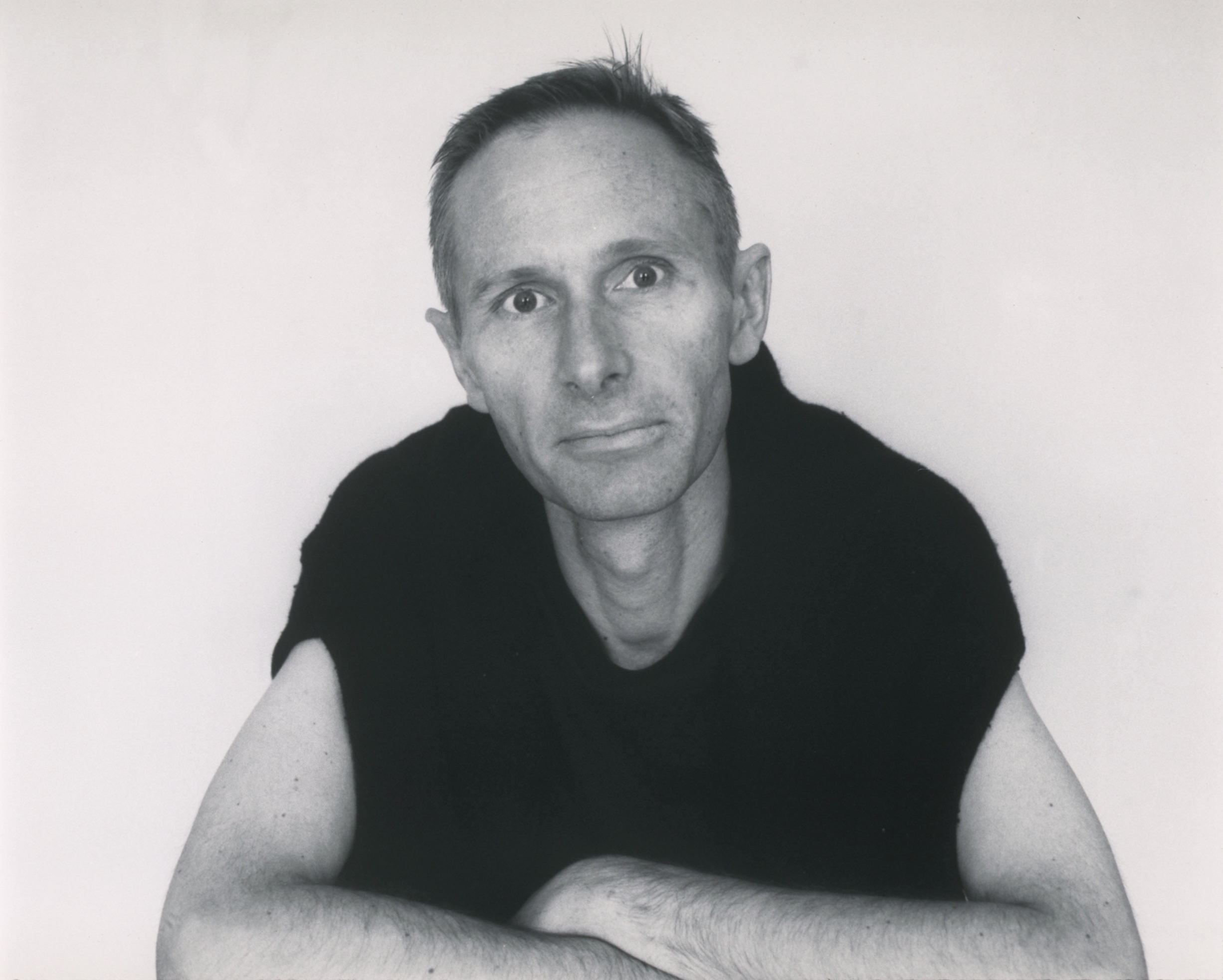 a black and white portrait of a man in his late thirties in a loose fitting
       black t-shirt resting both arms on (presumably) a table and looking directly at us