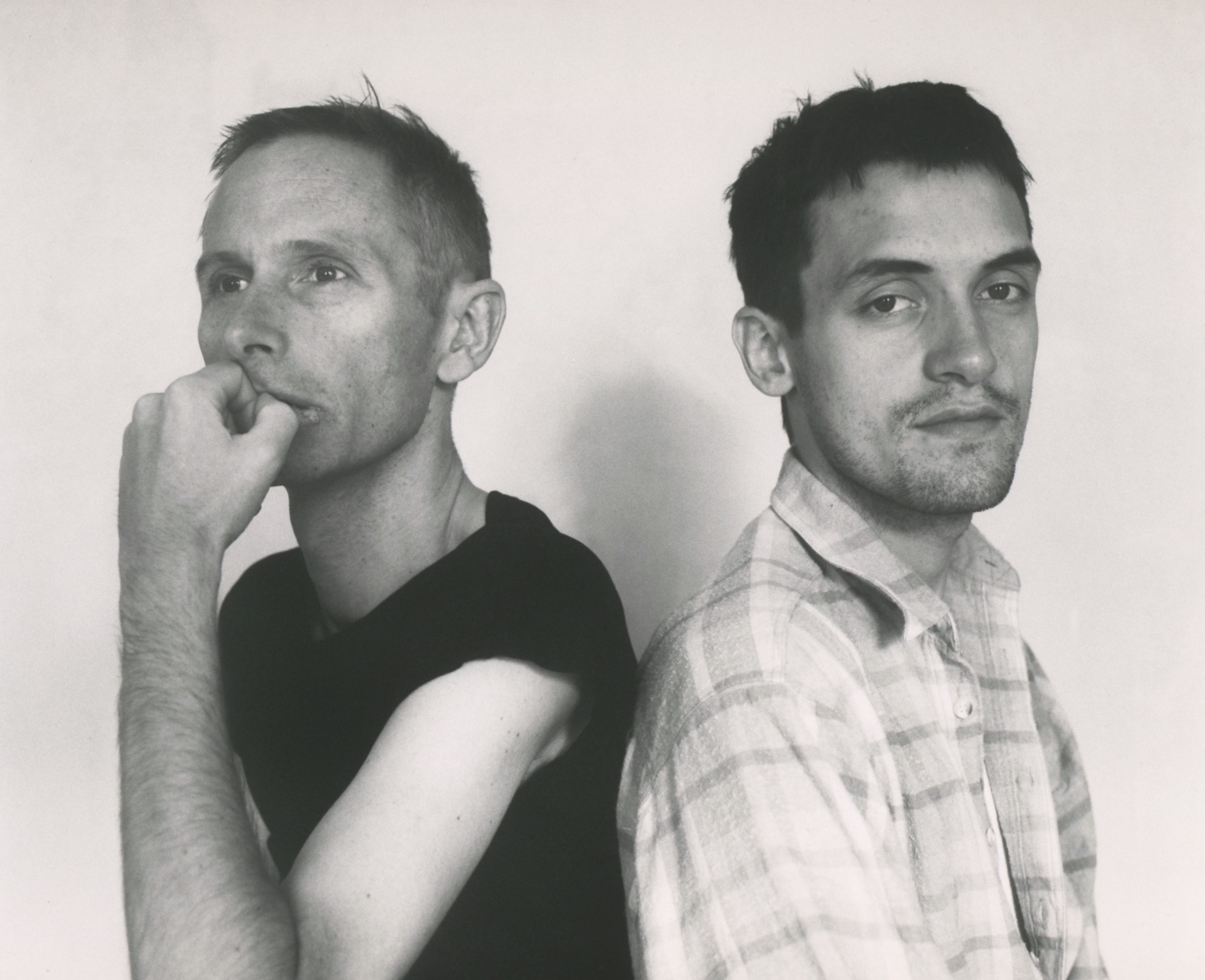 a black and white portrait of two men in their late thirties, sitting next
       to each other back to back, with one man looking at us and the other staring
       off into the distance