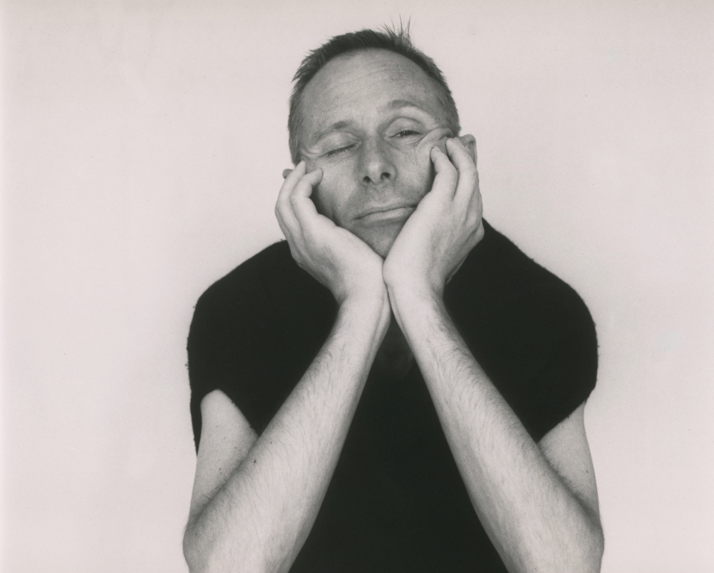 a black and white portrait of a man in his late thirties in a loose fitting
       black t-shirt cradling his head in his hands, one eye closed, the other
       peeking at us