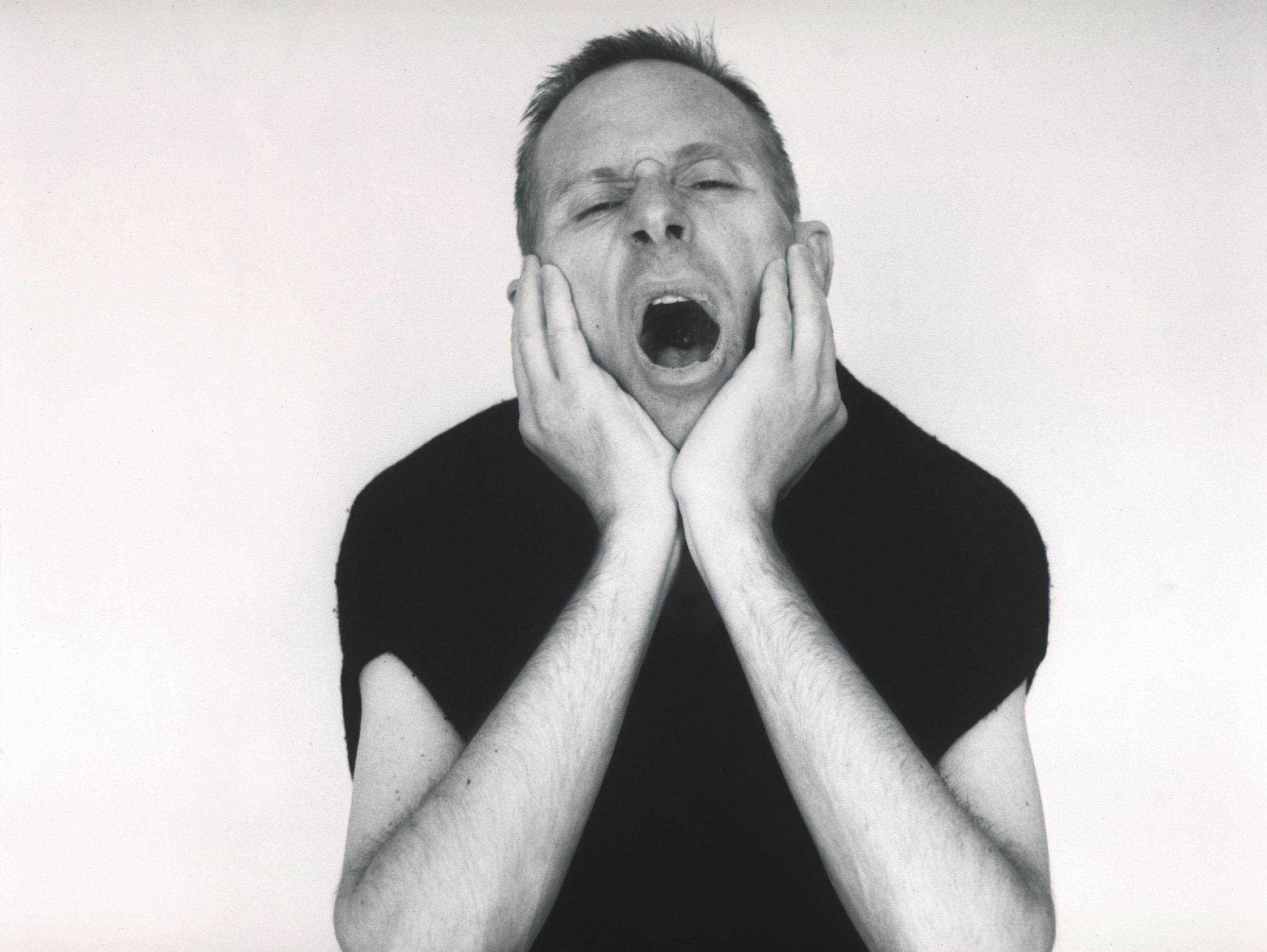 a black and white portrait of a man in his late thirties in a loose fitting
           black t-shirt cradling his head in his hands and yawning, mouth wide open