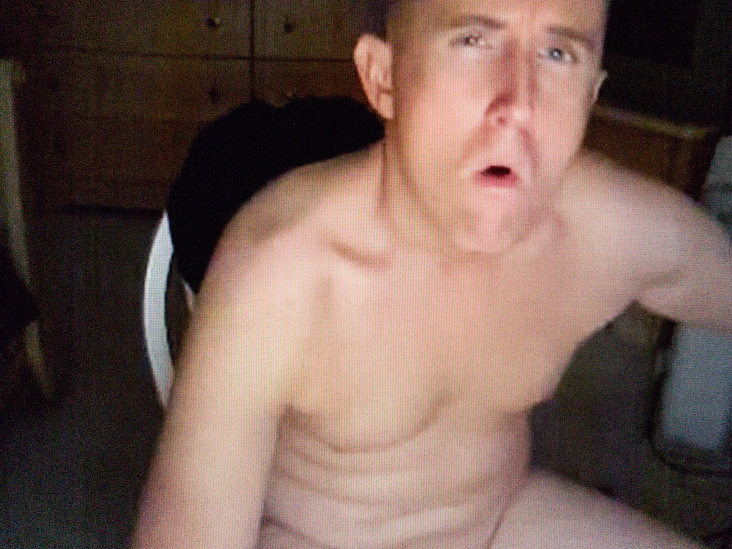 A buff, naked, young man sits on a chair in a darkened room. He looks directly into the camera and is opening his mouth in preparation for exhaling smoke.