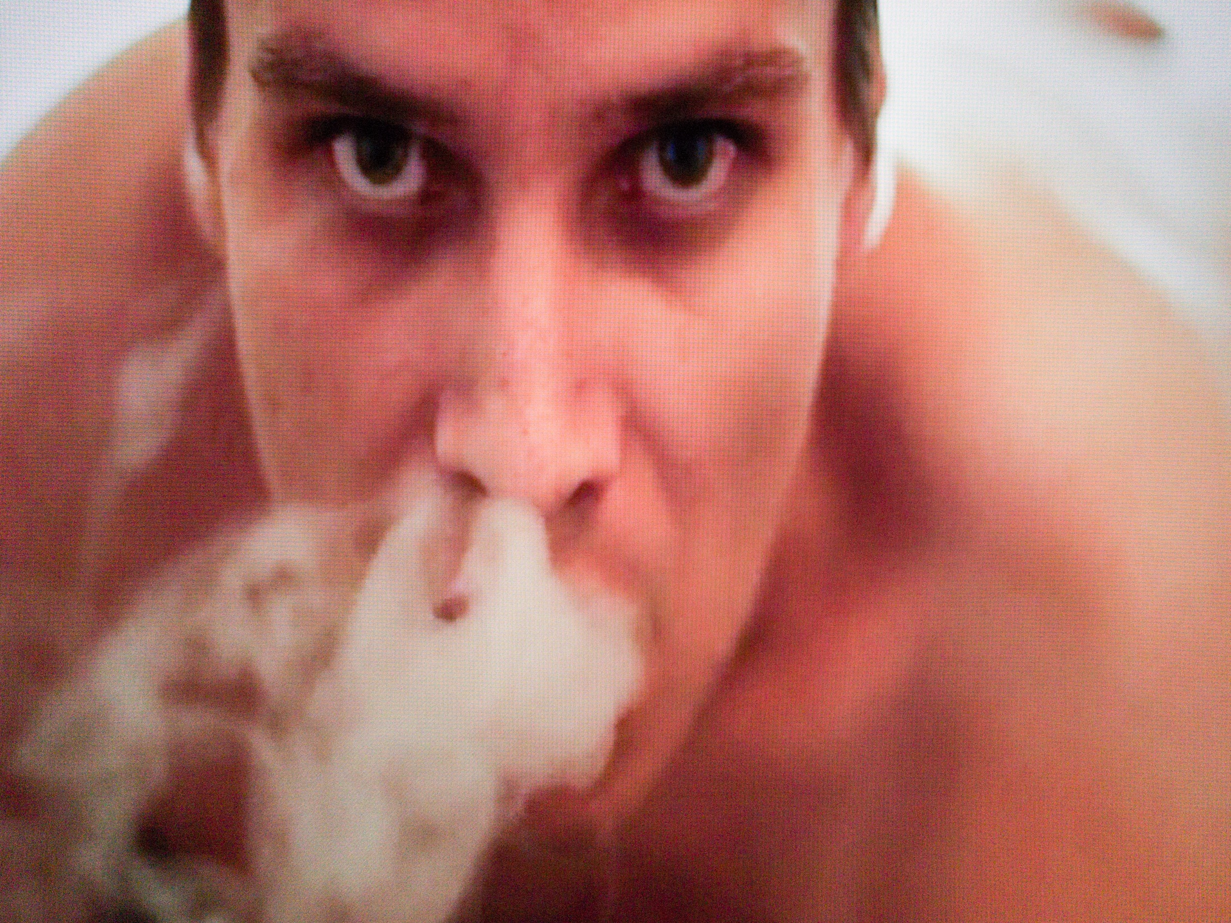 A fat, naked, young man lies on his stomach and looks directly at the camera, taking up the entire frame. He is exhaling smoke, which is drifting downward and covering his mouth and chin.