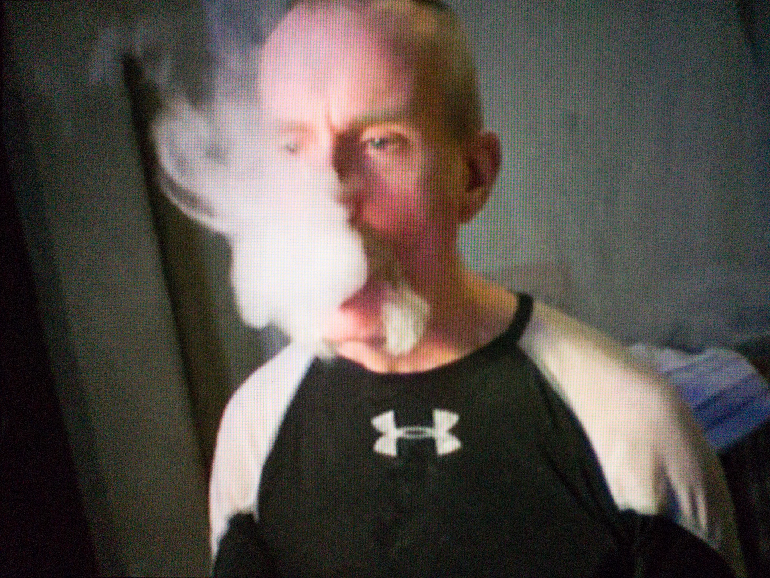 A middle-aged man with a grey horseshoe moustache and a black and white sweater sits in a darkened room. He is staring off into space to the side of the camera while blowing smoke, which is drifting upward and obscuring a third of his face.
