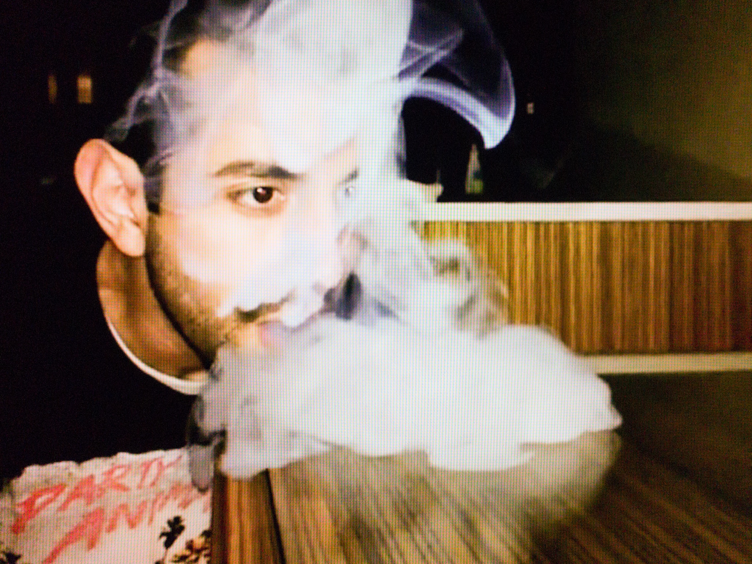 A scruffy young man wearing a ”party animal” t-shirt in a darkened room is slowly blowing smoke onto table, where it builds up.