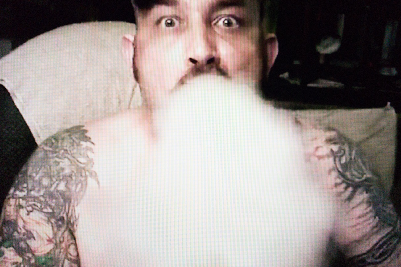 A bare-chested, bearded man with tattoos on arms and shoulders is looking straight at us with both eyes wide open, while exhaling a large volume of smoke that covers his entire chest