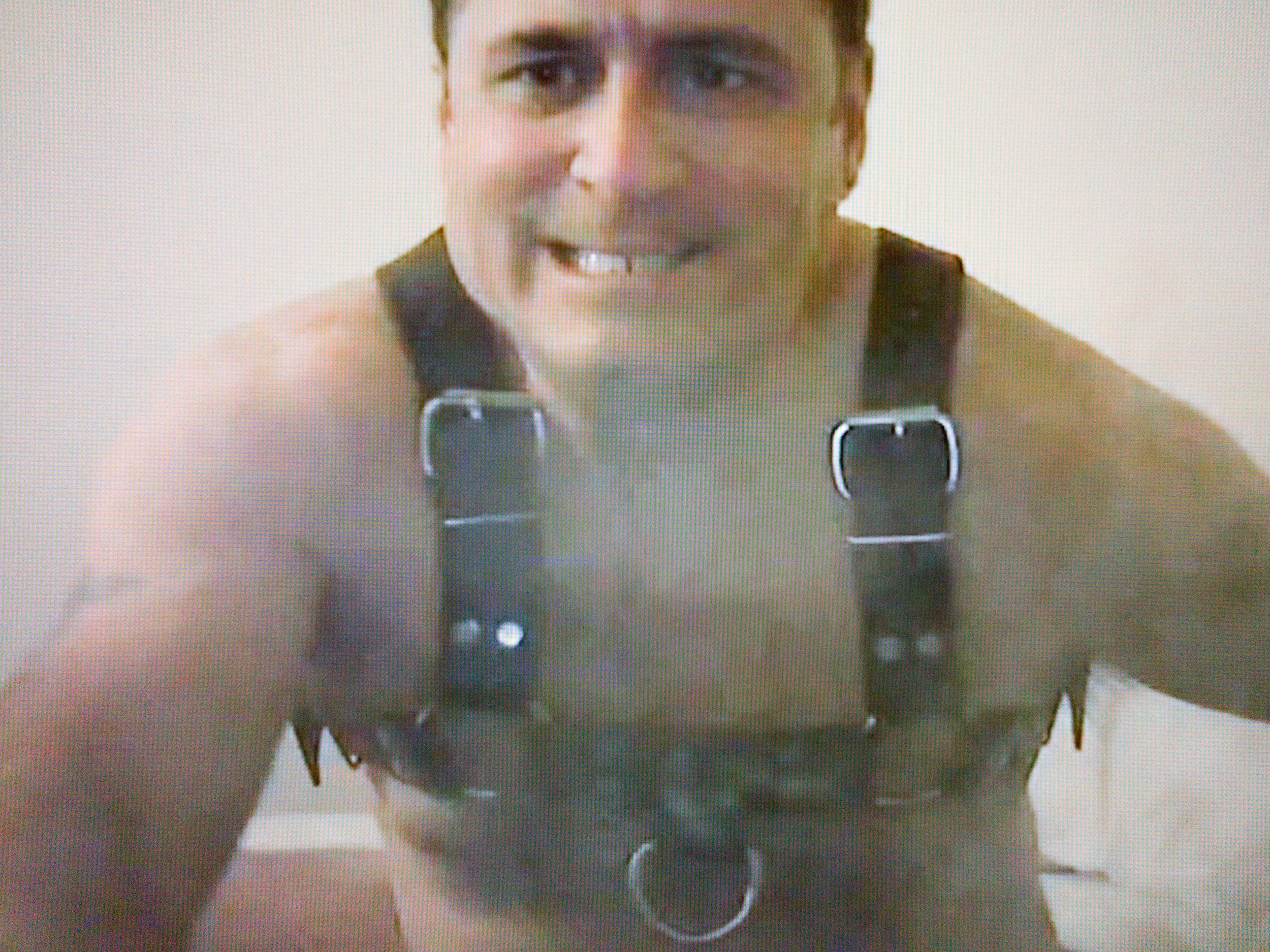 A naked man wearing a bulldog leather harness is leaning forward towards the camera, staring intently past the camera, and baring his teeth in a forced grin. There are traces of smoke in front of him all across the frame.