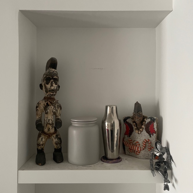 A white shelf in a white nook with two wooden ancestral spirits
             from Nigeria, a Cylon from the re-imagined Battlestar Galactica,
             a round grey cookie jar, and a cocktail shaker