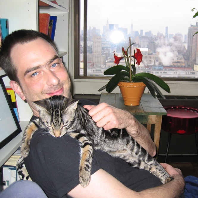 Robert looks into the camera with a happy smile; he is petting Esteban,
             who is stretched out napping across his chest, while his other arm is
             supporting Esteban